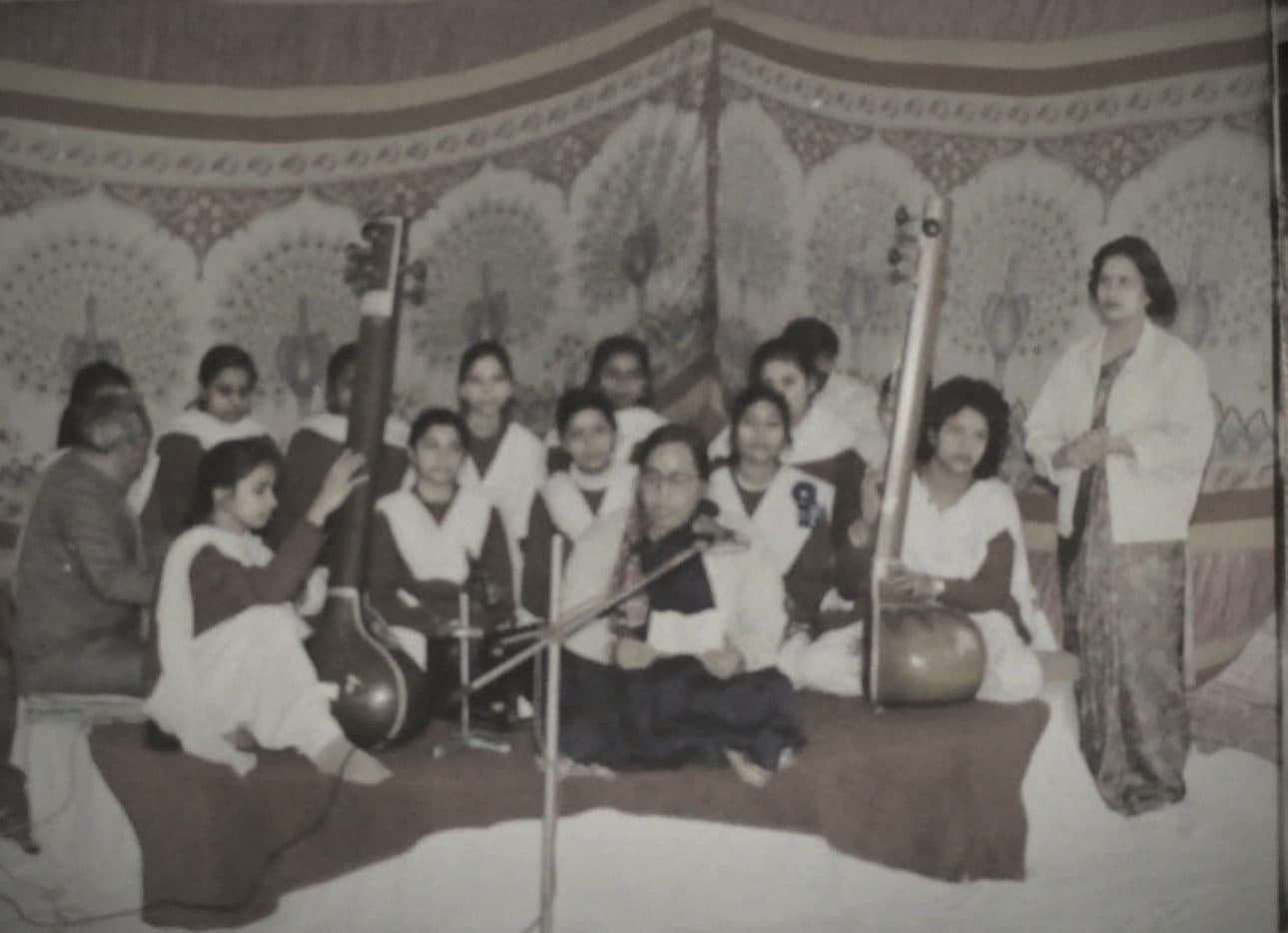 An archive photograph of a classical performance: two tanpuras upright, a harmonium at centre, and a full ensemble seated beneath a printed canopy
